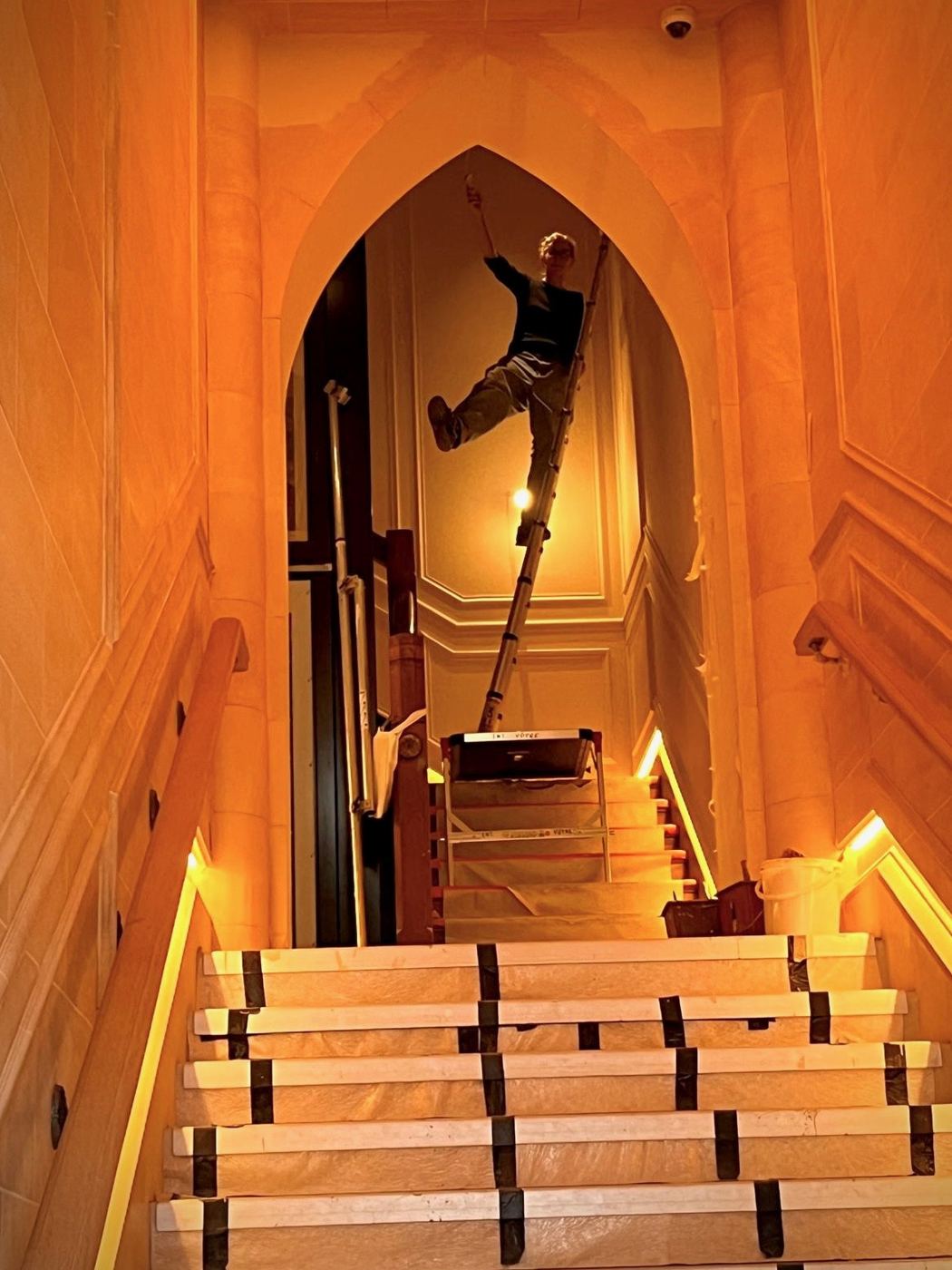 Emma sur une échelle sous une arche en ogive, dans une cage d'escalier en fausse pierre