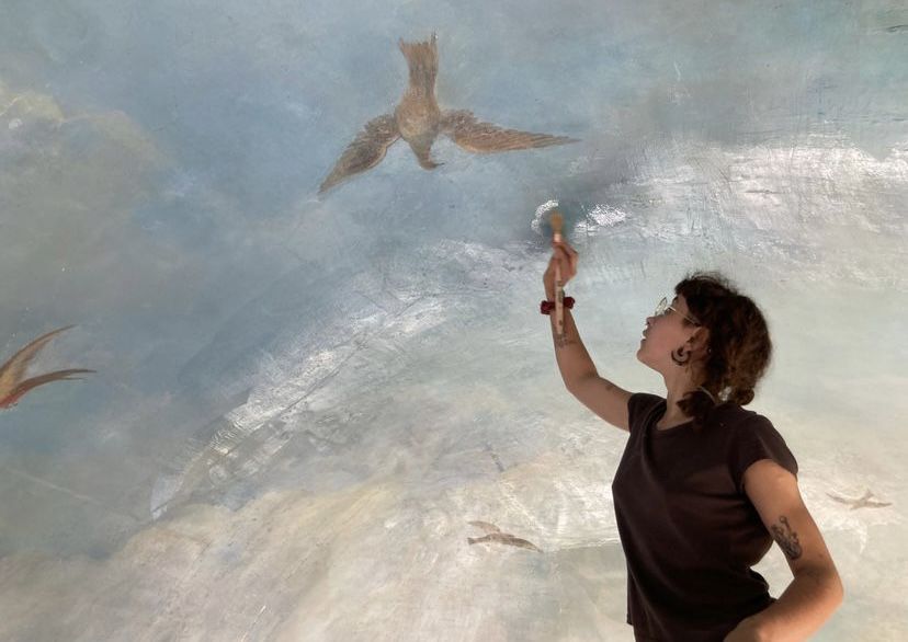Félicie, pinceau levé, retouche un plafond peint de ciel et d'oiseaux